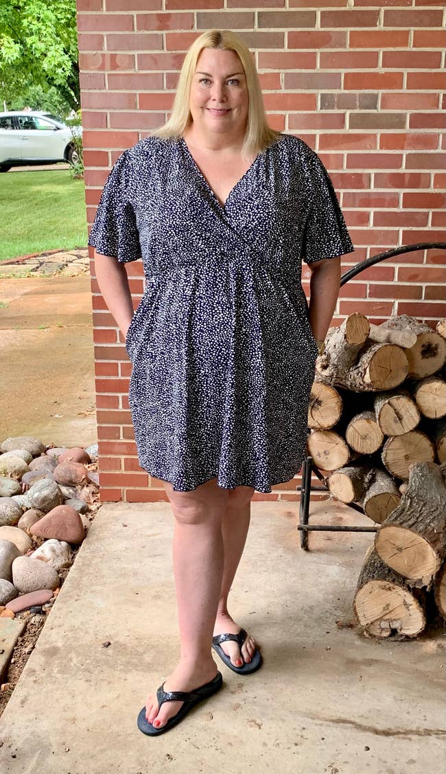 Person standing on a porch in a patterned summer dress and flip-flops, smiling near a woodpile