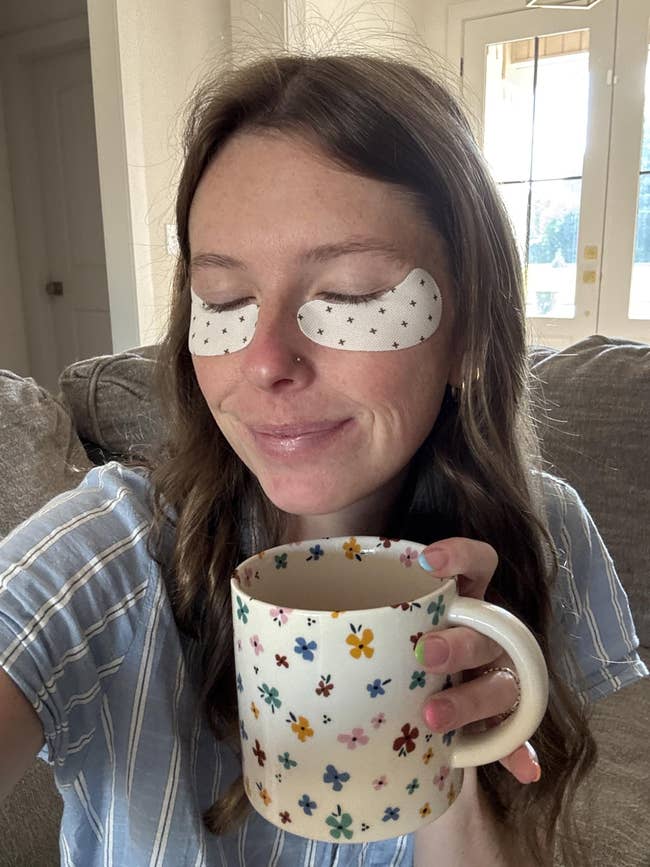 Person with eye patches smiling, holding a floral mug, wearing a striped shirt, sitting on a couch