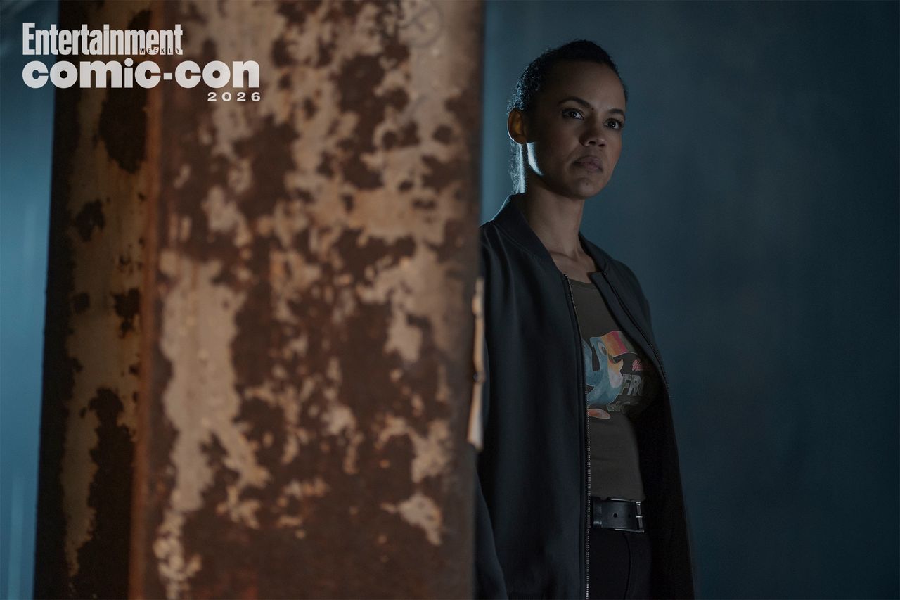 A person standing partially obscured by a rusted metal pillar with a stern expression under dim lighting