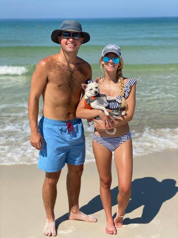reviewer wearing swim trunks on the beach, standing with a person holding a dog