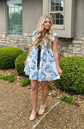 Person in a floral dress and heels stands outside a house with shrubbery