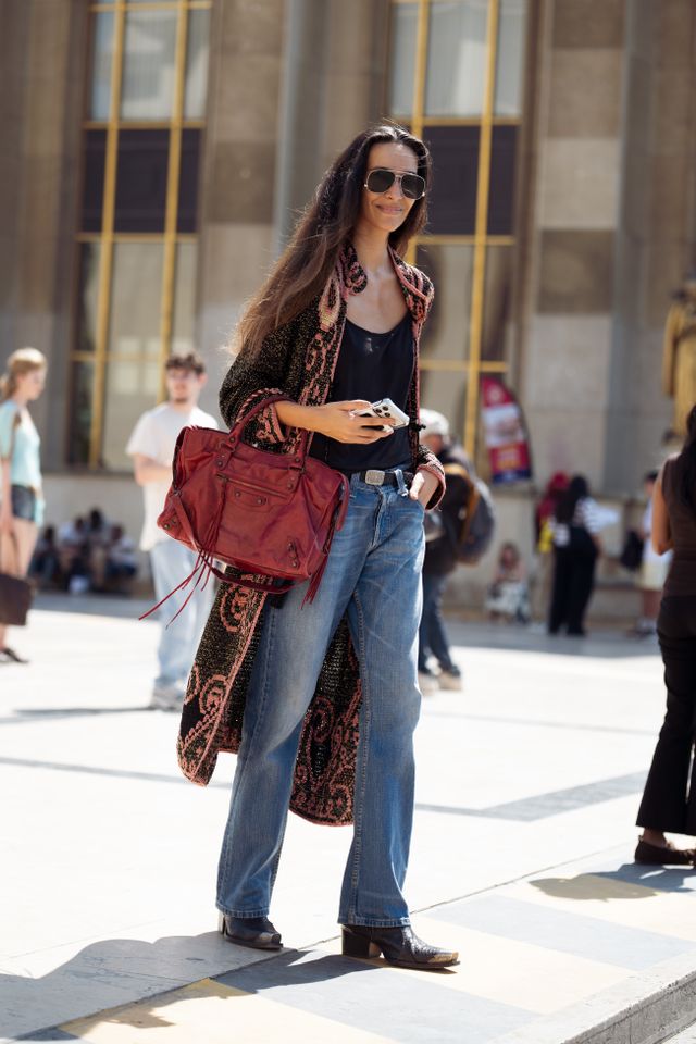 guest wears a black camisole, blue straight-leg jeans, a black and pink patterned long cardigan, oversized aviator sunglasses, carries a red Balenciaga City bag, holds a smartphone, and black pointed-toe ankle boots outside Elie Saab show