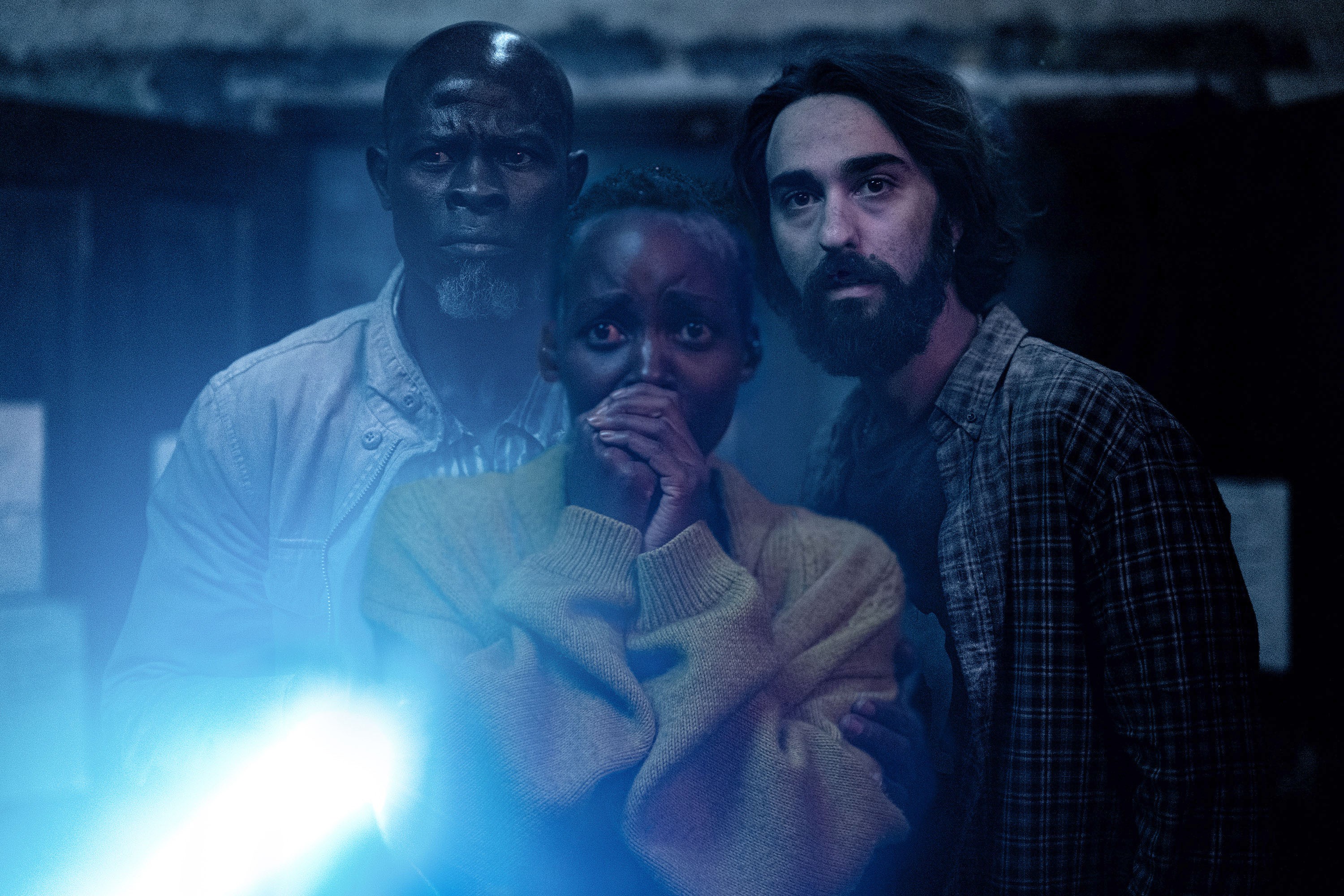 Djimon Hounsou, Lupita Nyong'o, and Alex Wolff look scared in a dark scene illuminated by a flashlight.