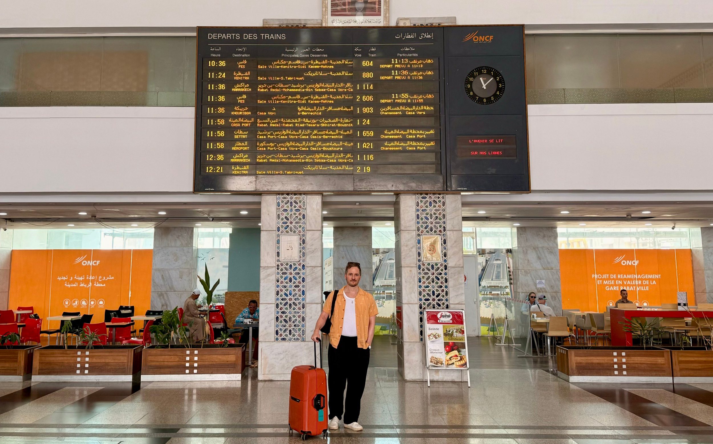 Matt Charlton standing with his suitcase in Rabat Ville station concourse, in front of an arrivals and departures information board