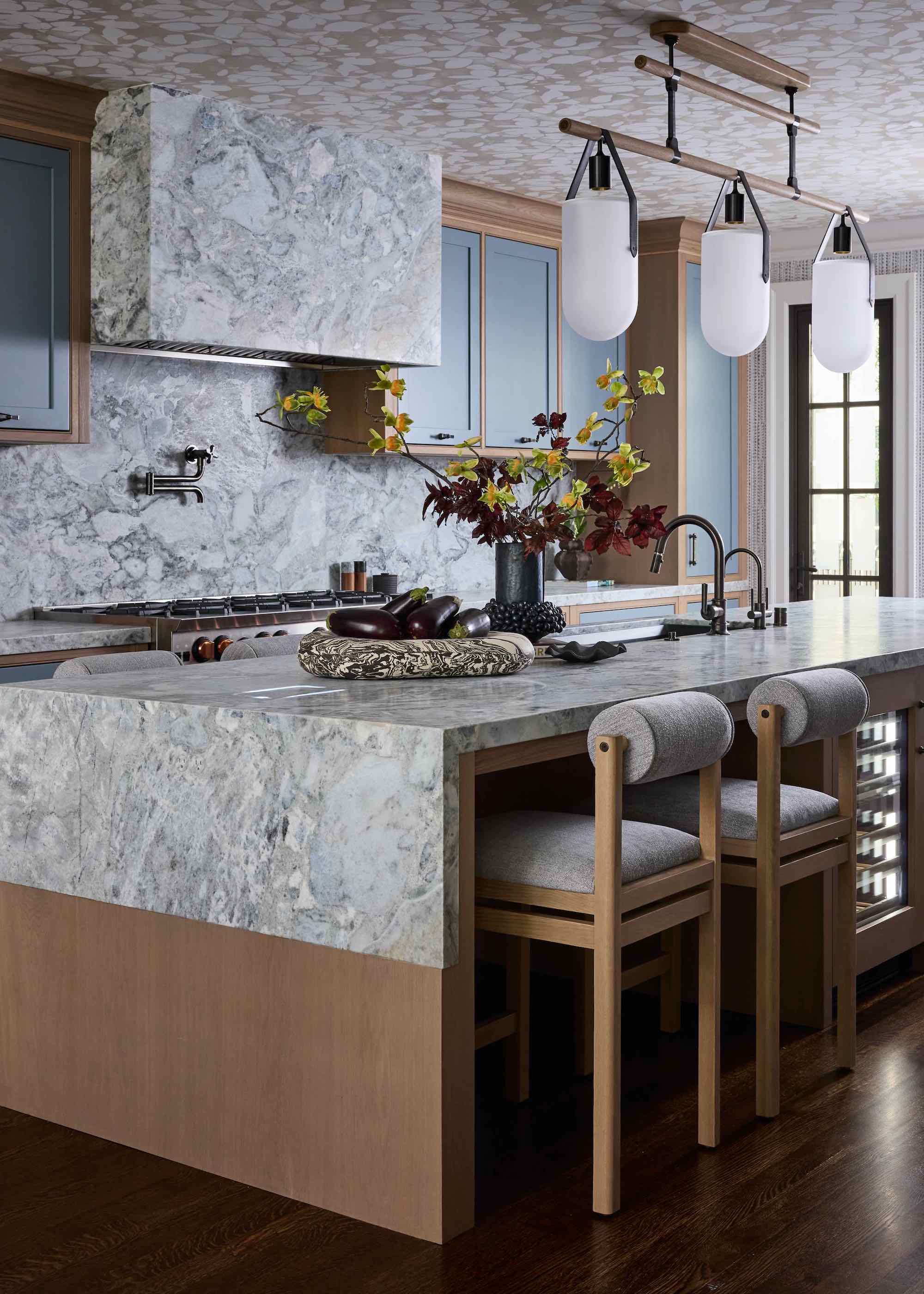 kitchen with wallpaper on ceiling, and grey swirling stone countertop, backsplash, and hood, with timber details, timber bar stools, three pendant lights, and vase of flowers