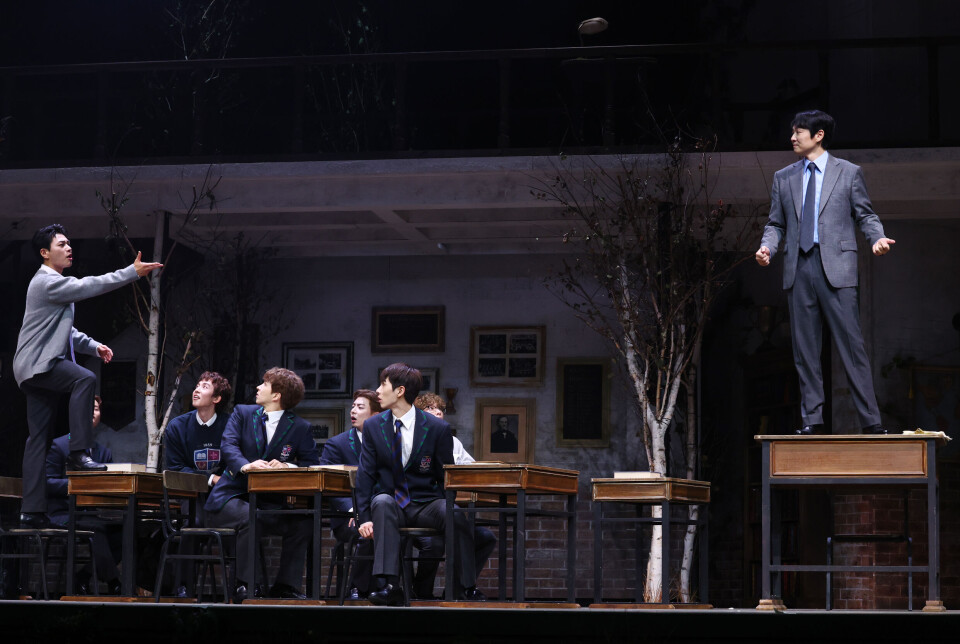 Actors in period costumes perform on a classroom-style stage during a play press call.