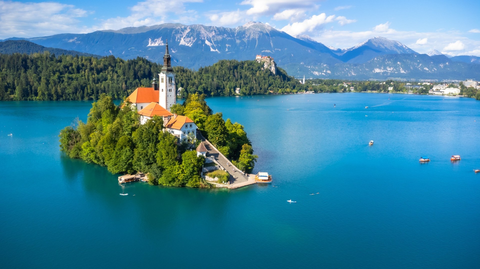An aerial view of Lake Bled's island surrounded by blue water, with mountains in the distance.
