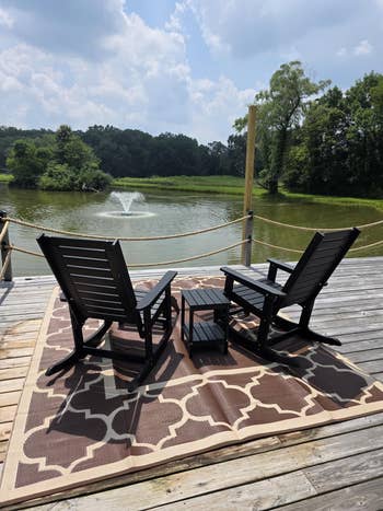 Two wooden chairs and a table on a patio overlooking a pond with a fountain, surrounded by trees. Ideal setting for outdoor furniture shopping