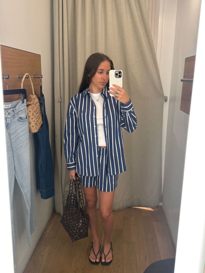 A person in a fitting room wearing a striped outfit and holding a patterned bag taking a selfie in a mirror