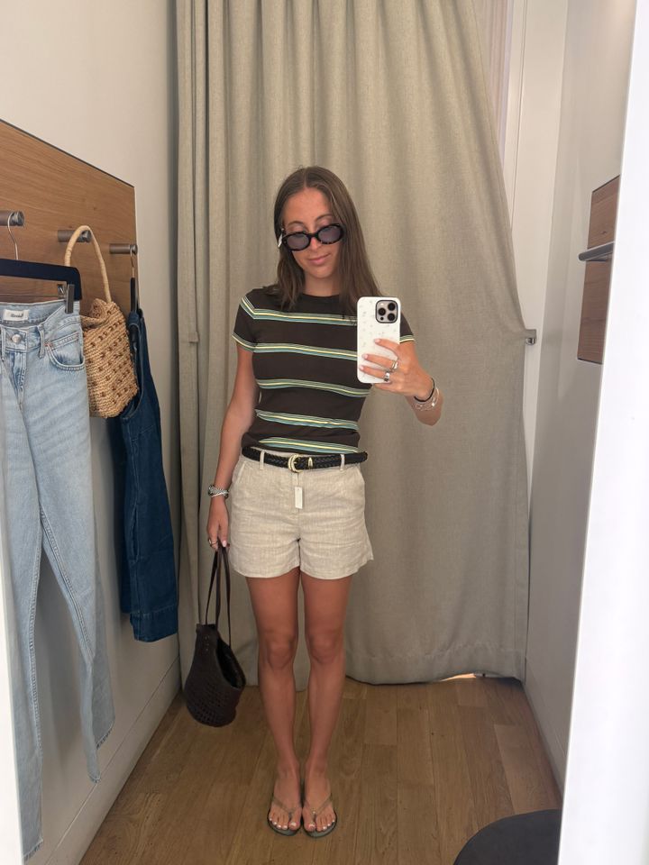A person taking a mirror selfie in a fitting room wearing a striped shirt shorts and sunglasses holding a black bag and a phone