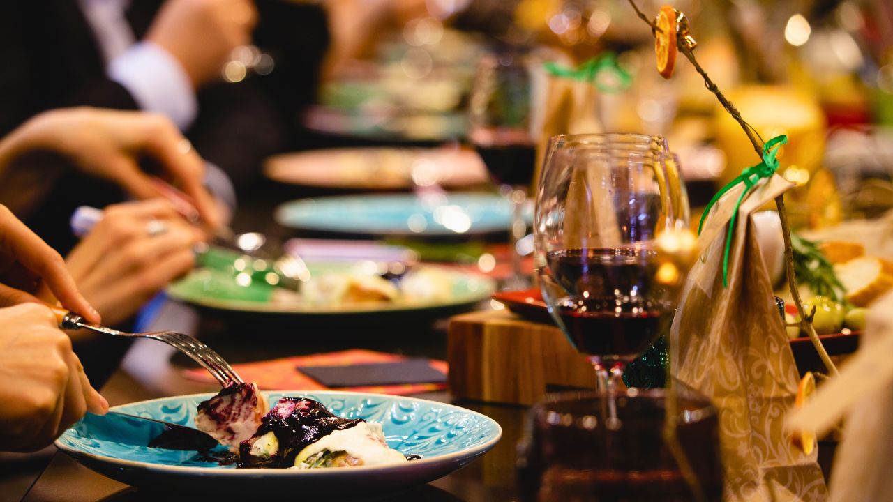 Hands eating a berry-topped dessert on a blue plate beside a glass of red wine.