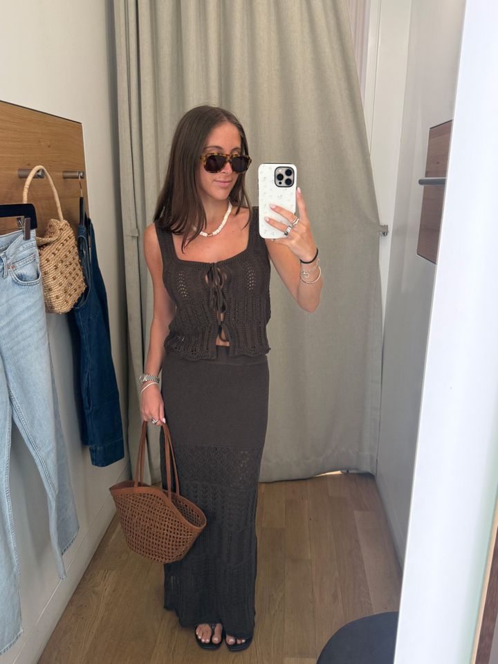 A woman wearing a sleeveless top and long skirt taking a selfie in a fitting room holding a basketstyle bag