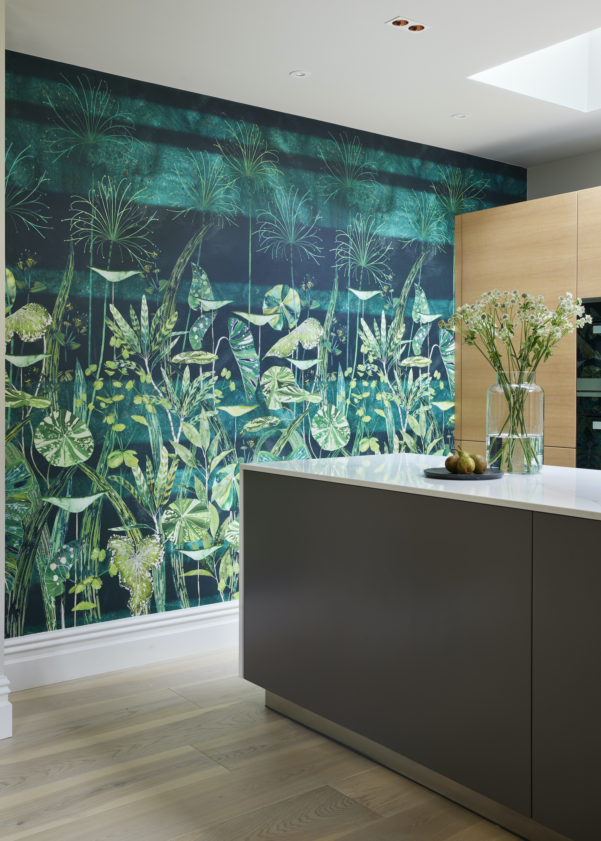 kitchen with leafy mural wallpaper, with blonde timber floorboards, timber cabinetry, and gray kitchen countertop with white stone countertop with vase of flowers and skylight