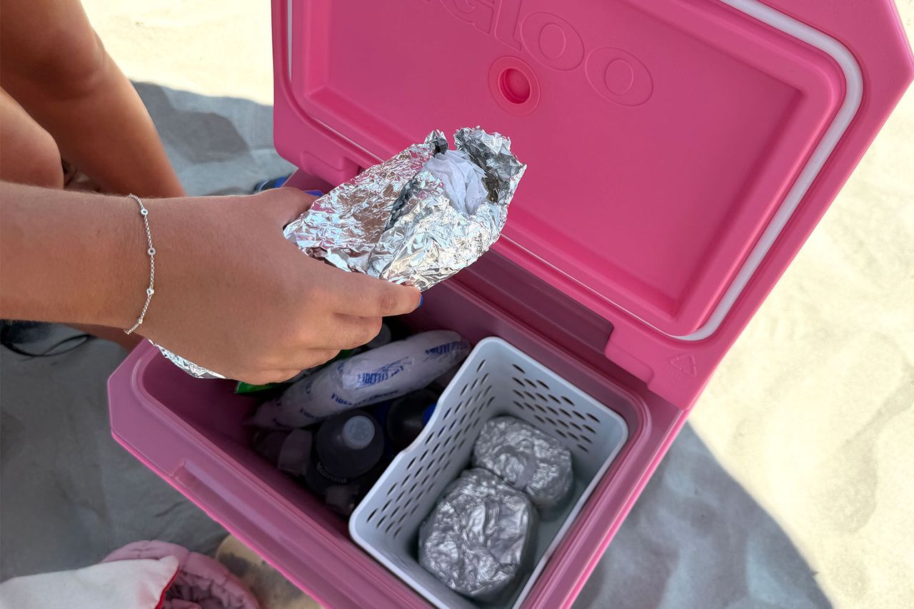 A pink cooler on the ground with food items like foilwrapped packages and bottles inside and a persons hand placing an item in it
