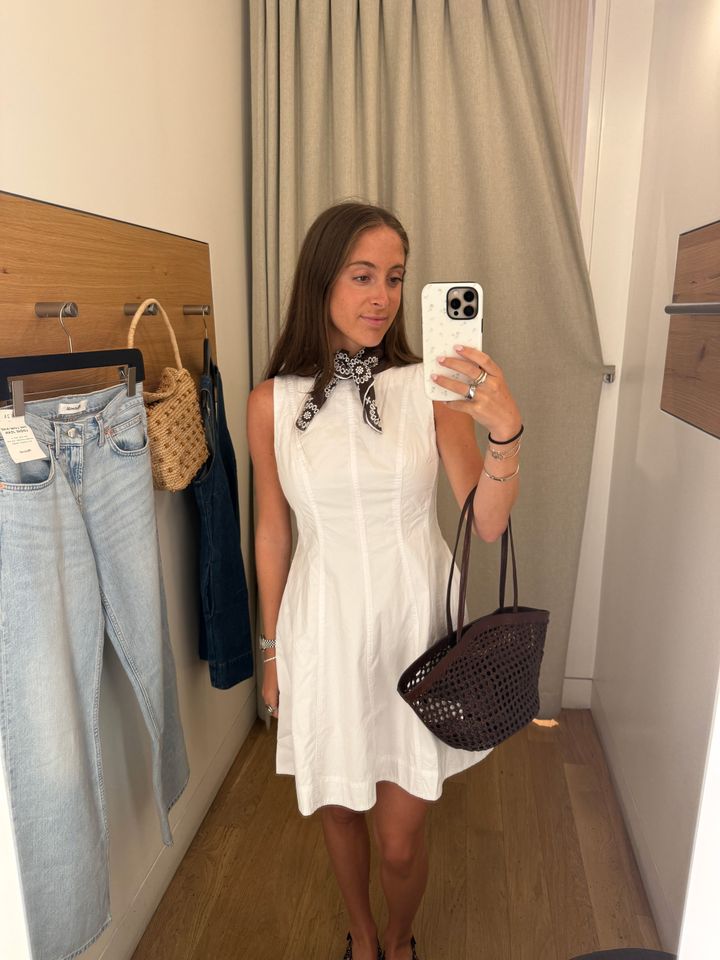 Person in a changing room taking a mirror selfie wearing a sleeveless white dress with a scarf holding a bag