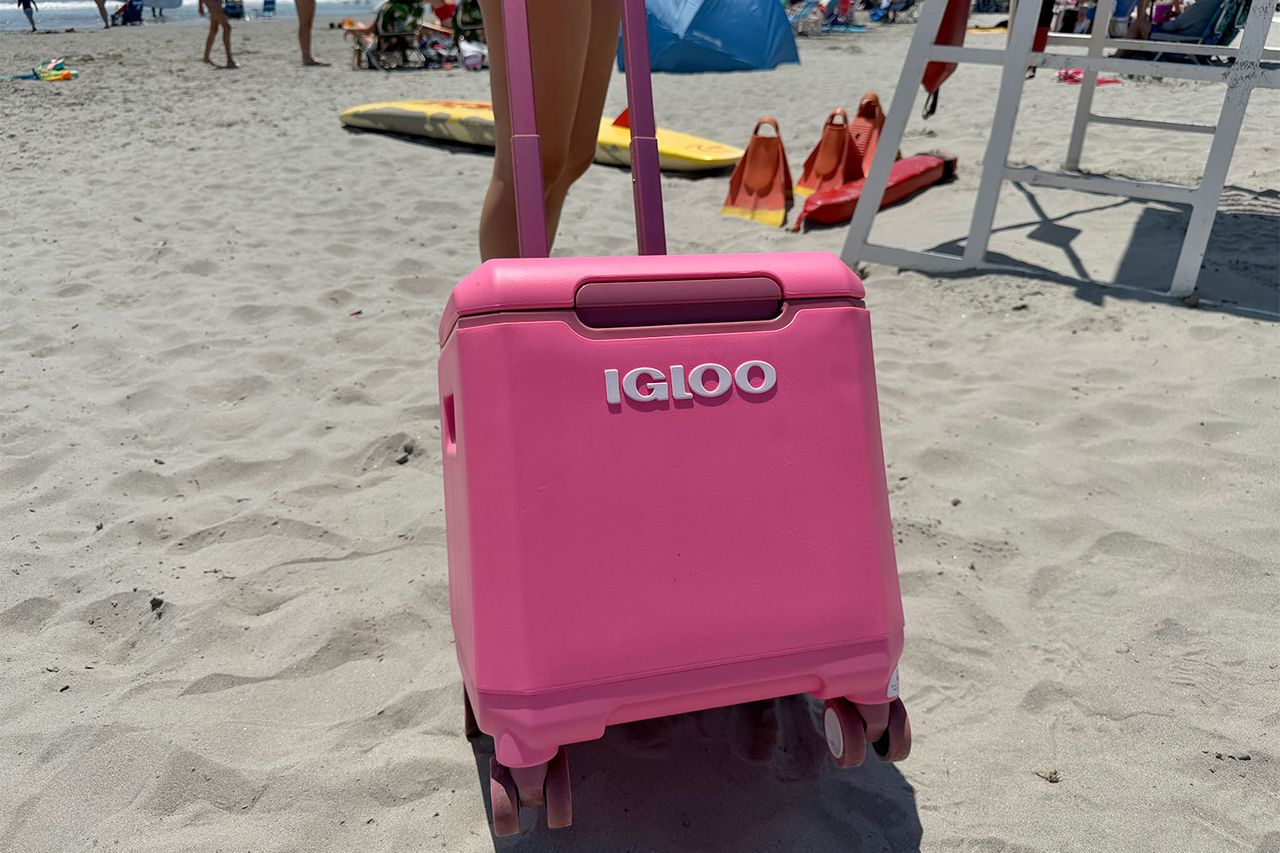 A rolling Igloo cooler positioned on a sandy beach near surfboards and beachgoers