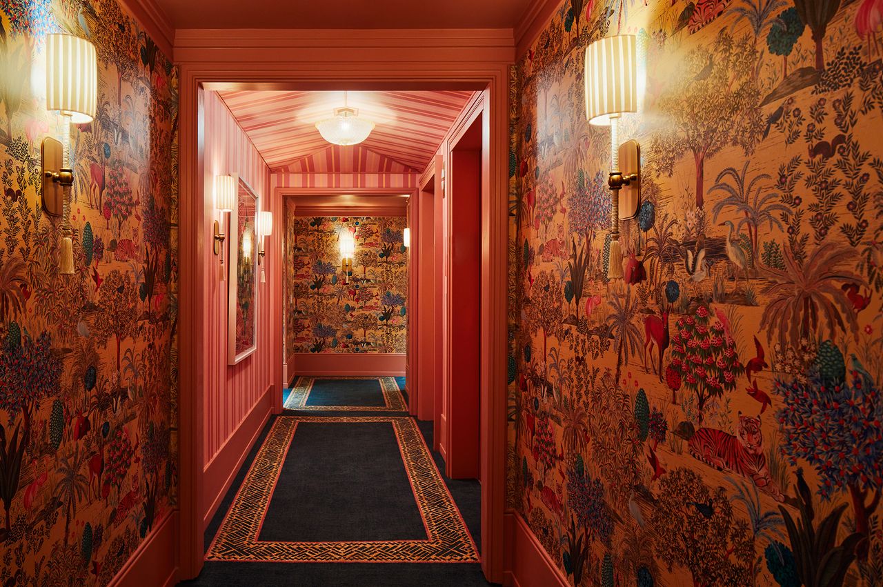 A decorative hotel hallway with patterned wallpaper wall sconces and a dark carpeted walkway leading to a lit area
