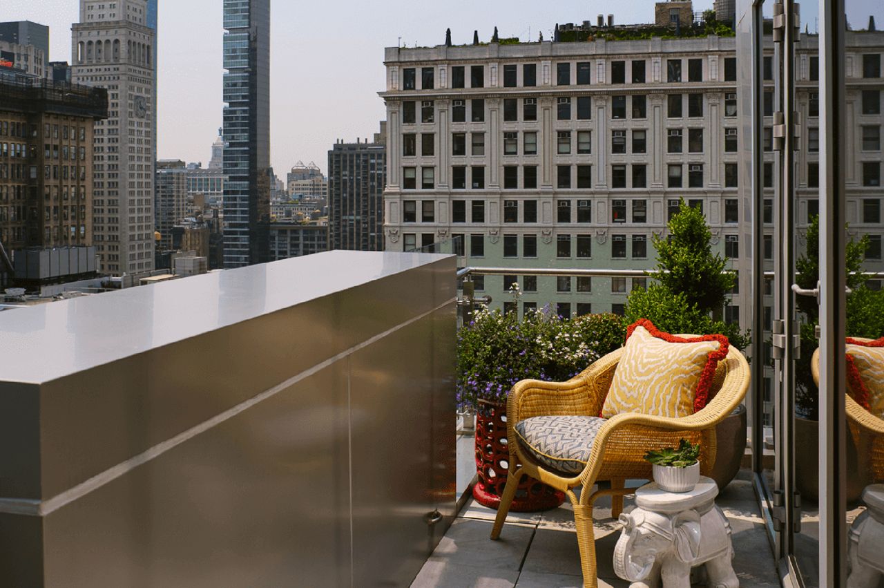 A rooftop terrace featuring a minimalist railing wicker armchair small side table potted plants overlooking a cityscape with tall buildings