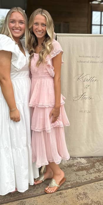 Two people in ruffled dresses standing beside a sign at a rehearsal dinner, with outdoor venue seating in the background