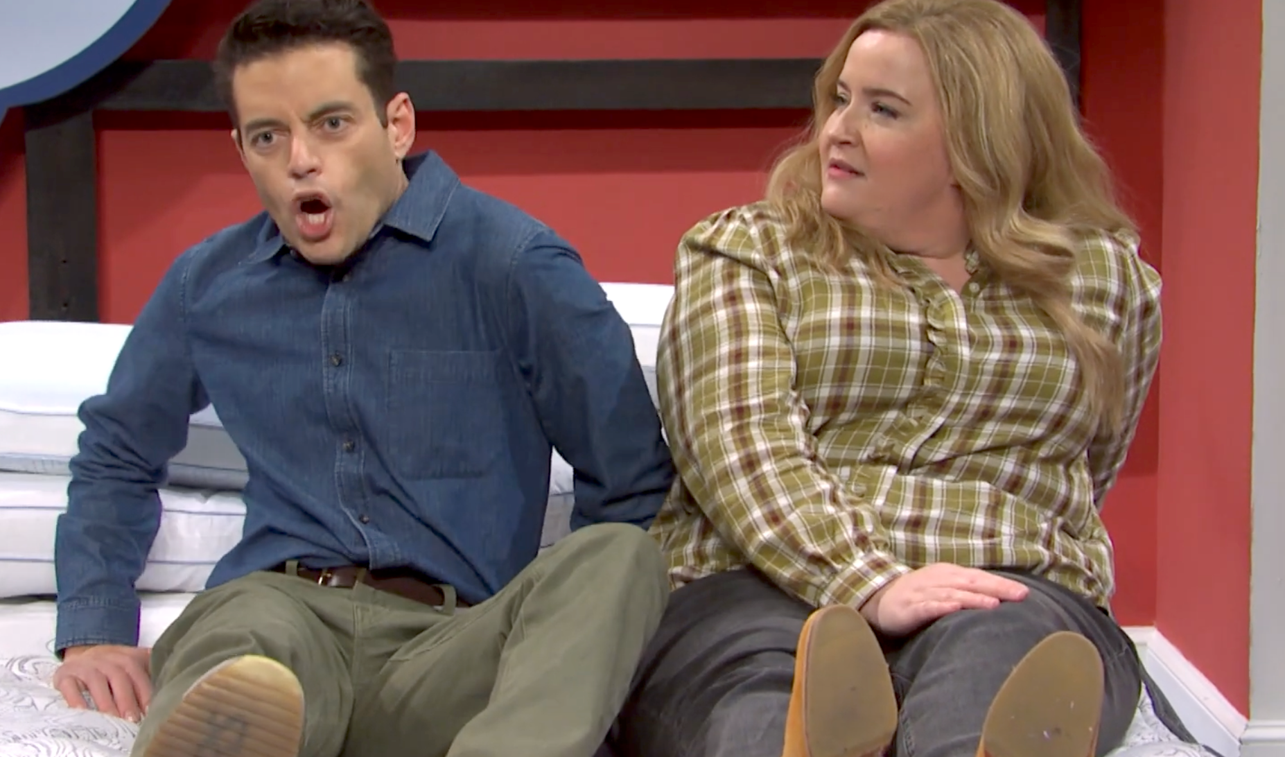 Two people sit on a bed. The person on the left looks surprised, and the person on the right is smiling, wearing casual attire