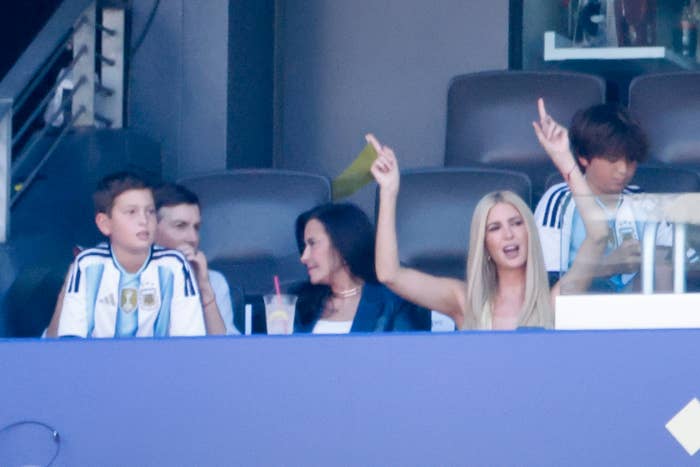 People are watching a sports event from a private box. One woman is cheering with both arms raised