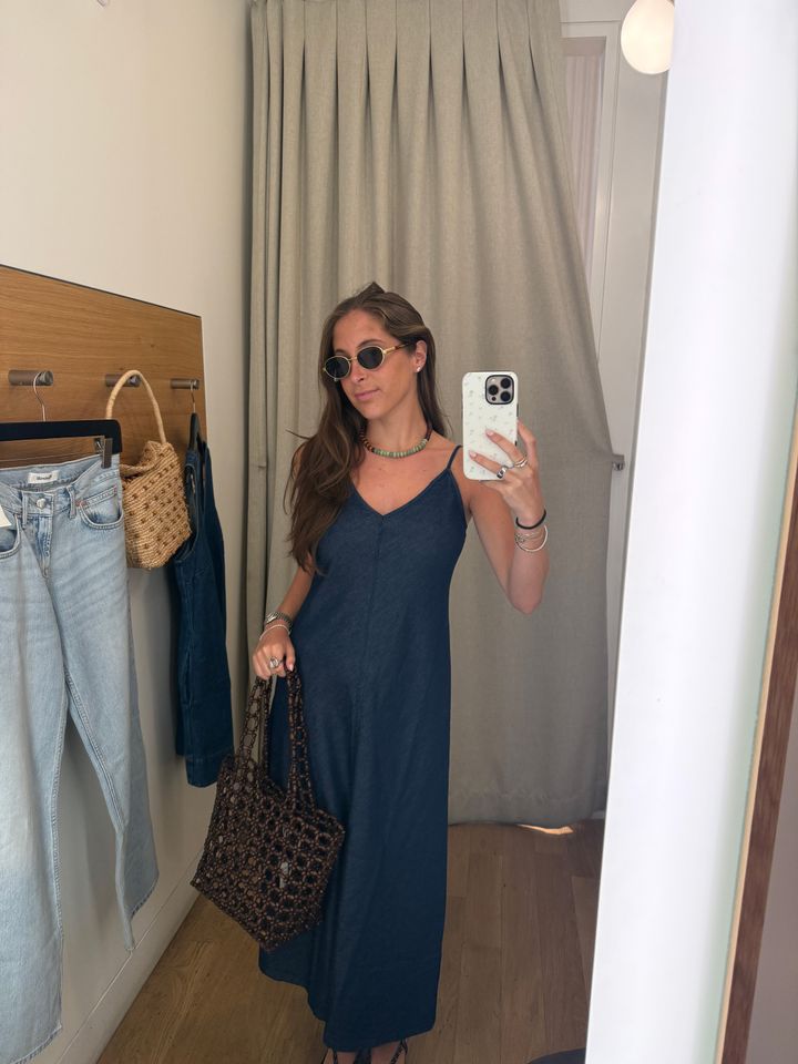 Person posing in a retail dressing room with a summer outfit holding a phone and wickerstyle bag