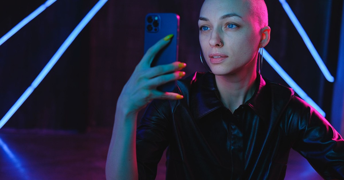 Young bald female in black shirt taking pictures on mobile phone while standing in neon lights in dark studio