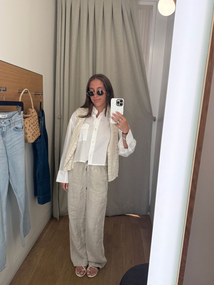 Person taking a mirror selfie in a fitting room wearing a light outfit and sunglasses with clothing items hanging in the background