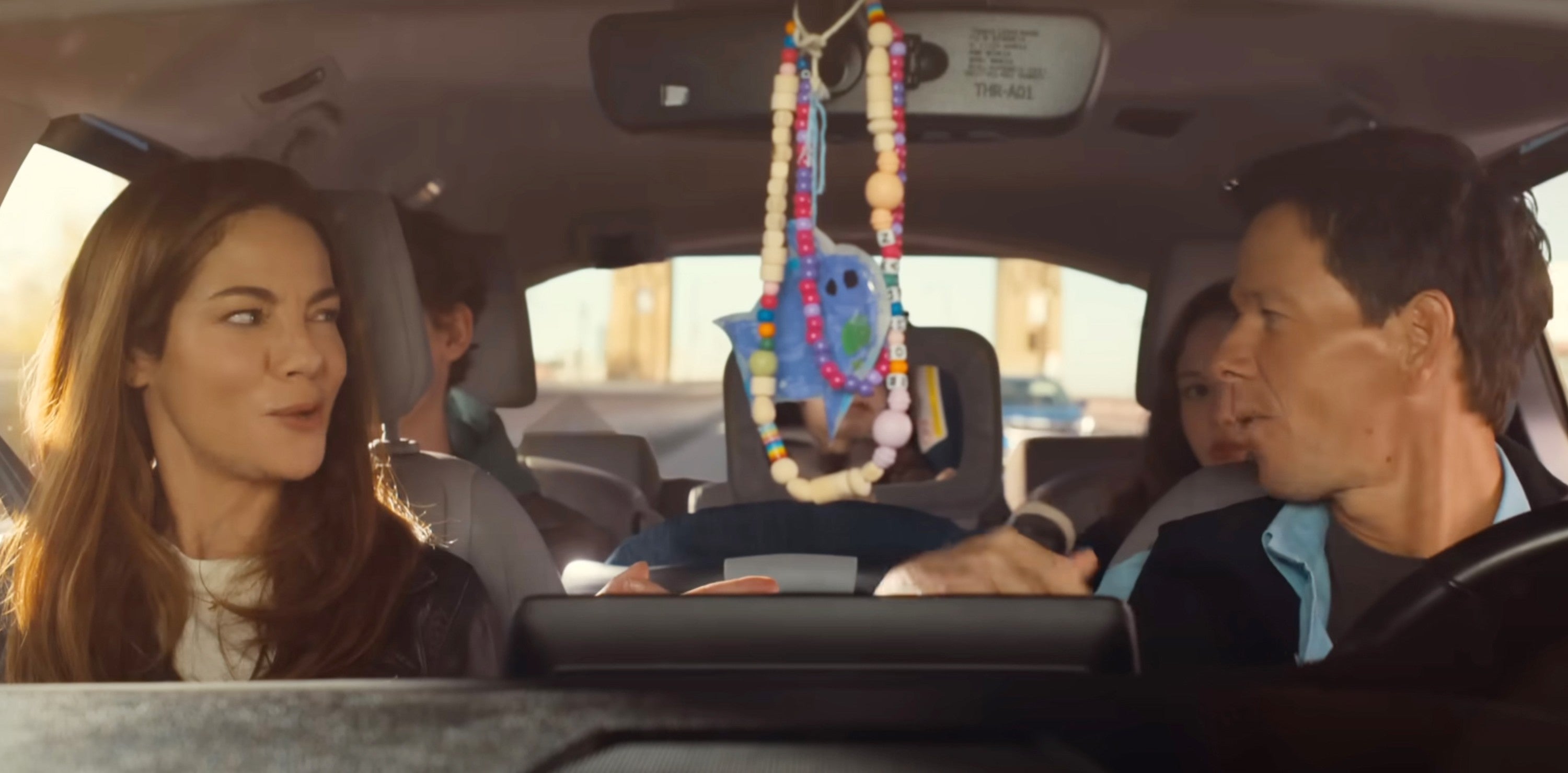A couple in a car share a playful moment, with a child secured in a car seat behind them, and colorful toys hanging from the rearview mirror