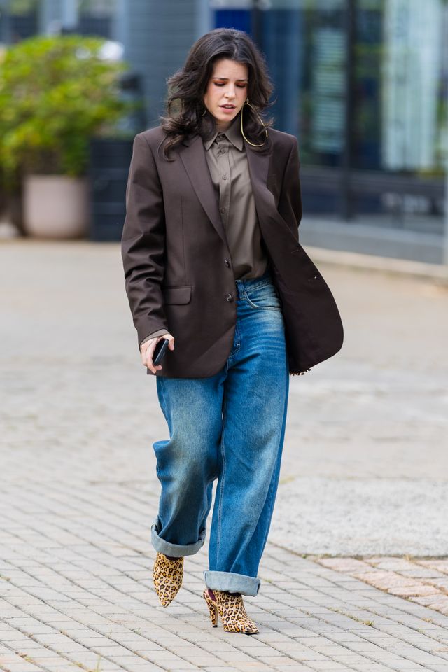 A guest wears medium-length wavy dark brown hair, olive khaki button-down shirt, dark chocolate brown blazer, structured shoulders, notched lapels, light wash blue straight-leg jeans, cuffed hem, leopard print pointed-toe high heels, gold hoop earrings, black smartphone in hand, bracelet on wrist before Mondepars fashion show