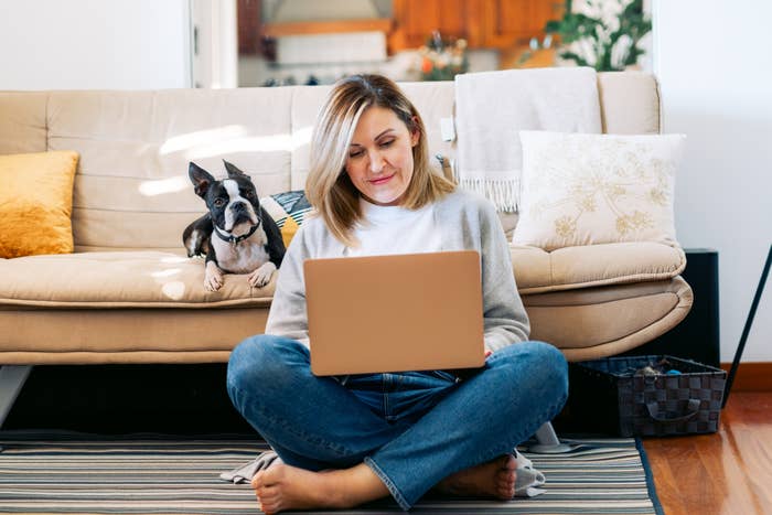 Woman sitting cross-legged on the floor, using a laptop, with a small dog on the couch behind her. Cozy, casual living room setting