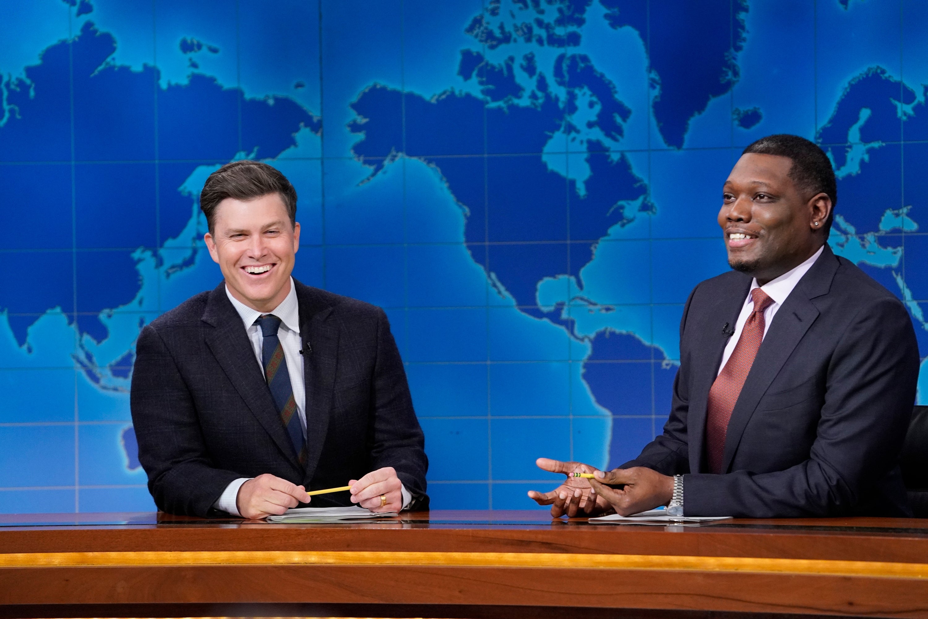 Two people in suits sit at a news desk, smiling and holding pencils, with a world map displayed in the background