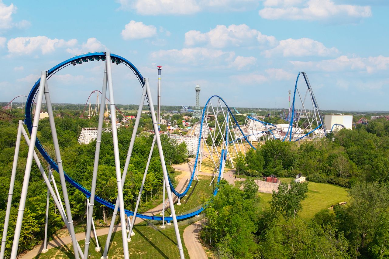 View of a theme park with a roller coaster spanning a wooded area and buildings in the distance