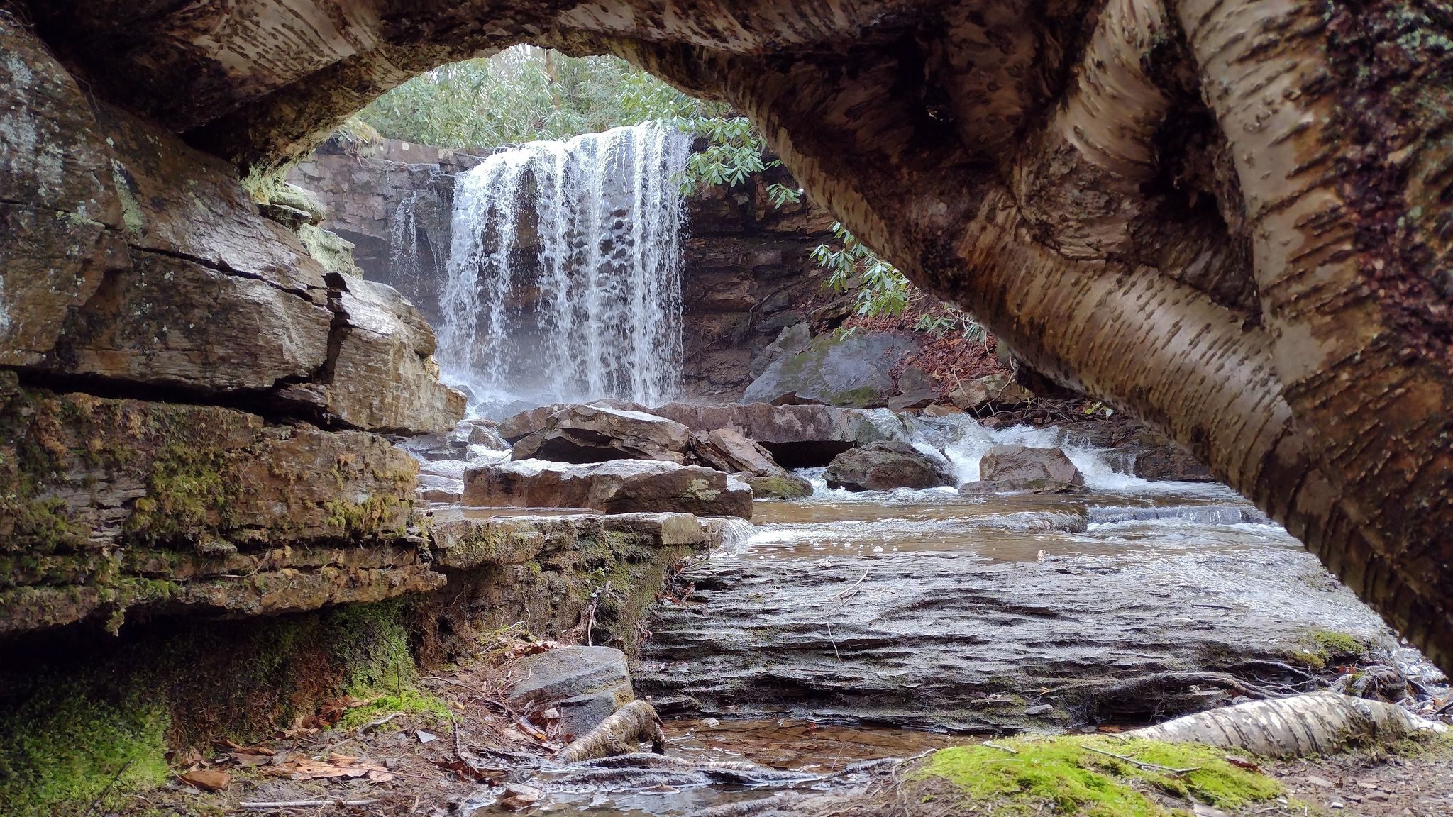 A natural arch frames a beautiful waterfall, along Cole Run Trail in Pennsylvania, surrounded by tiered rocks.