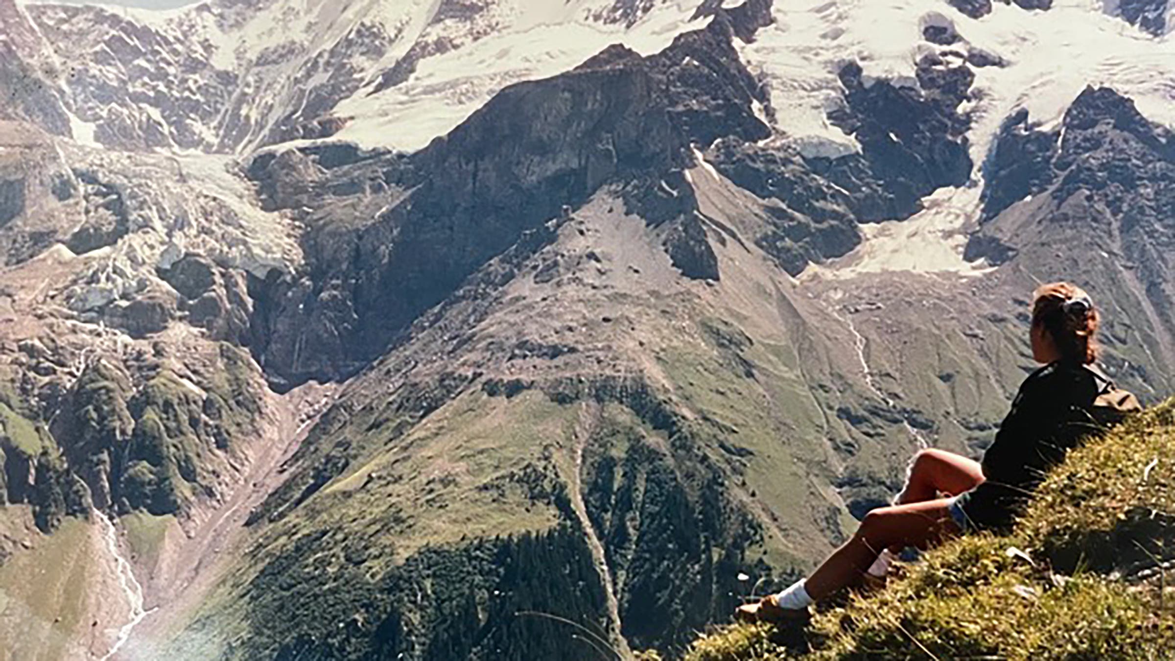 Woman sitting on mountainside enjoying view across a valley to a towering range in the distance