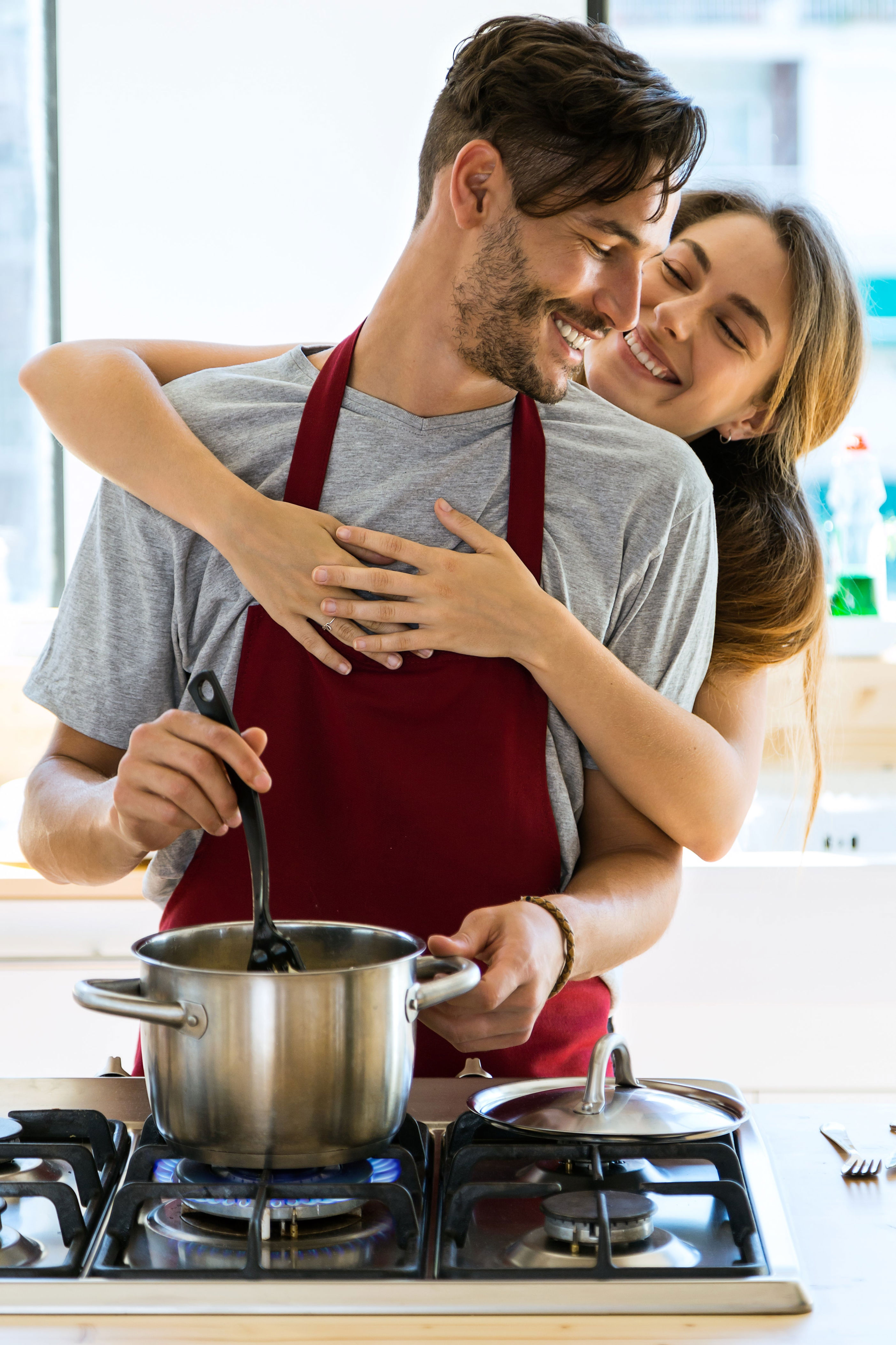Couple cooking together, smiling as one stirs a pot on the stove, both wearing casual clothing; a joyful kitchen moment