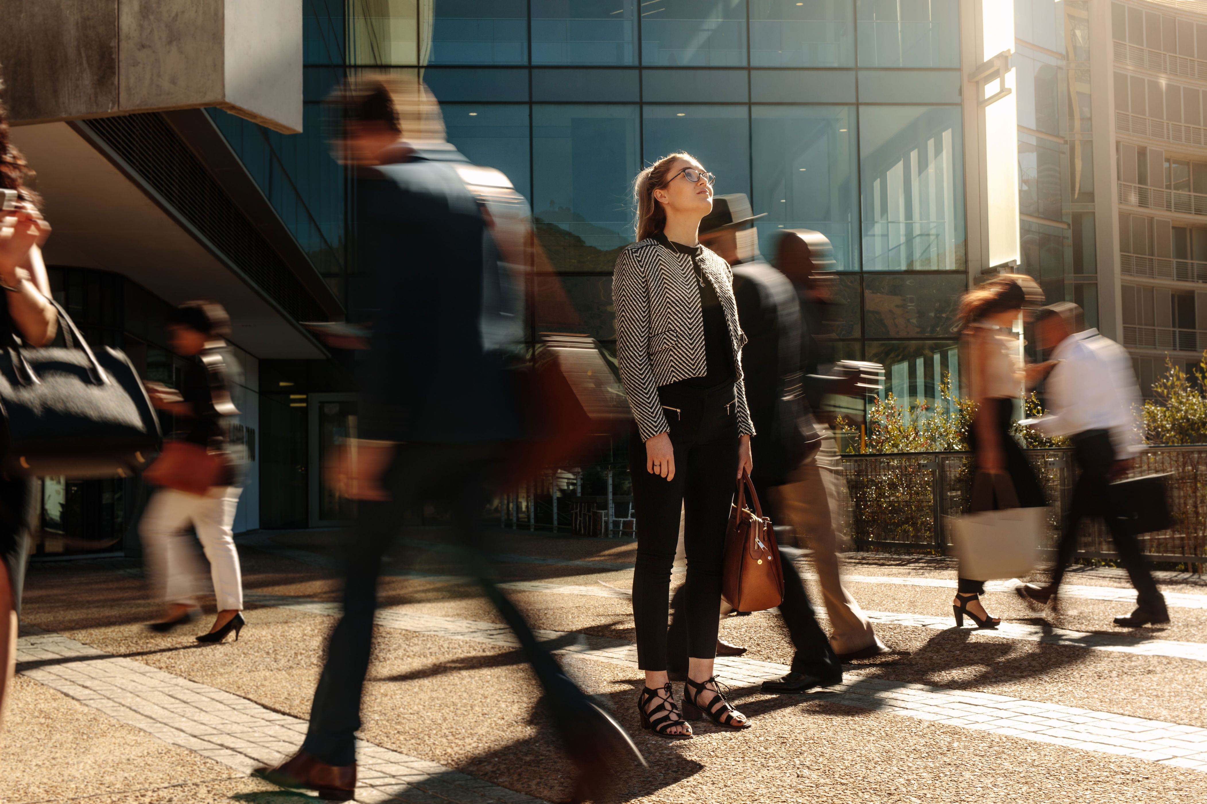 A woman stands still in a busy urban setting as people rush past her, creating a blur effect, suggesting a theme of mindfulness or contemplation
