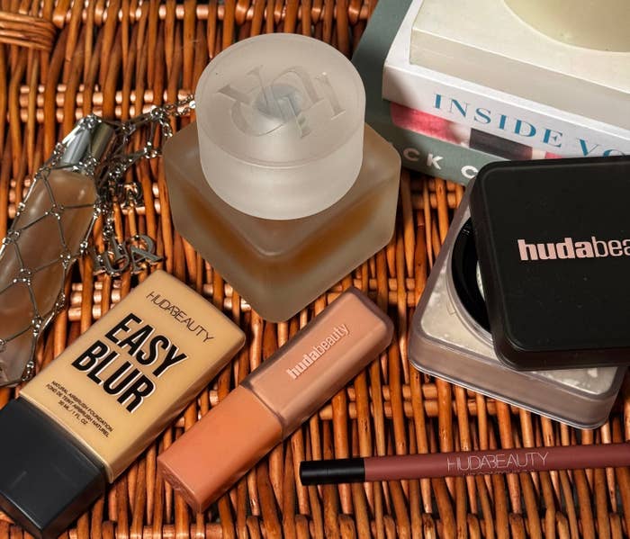 Beauty products arranged on a wicker surface, including foundation, concealer, lip gloss, a compact, and a makeup brush with branded labels