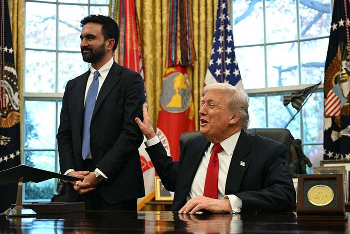 Two men in a formal setting, one seated with an expressive gesture, the other standing and smiling, flags displayed in the background