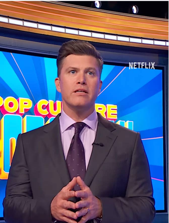 A person in a suit stands in front of a "Pop Culture" game show set with a concentrated expression