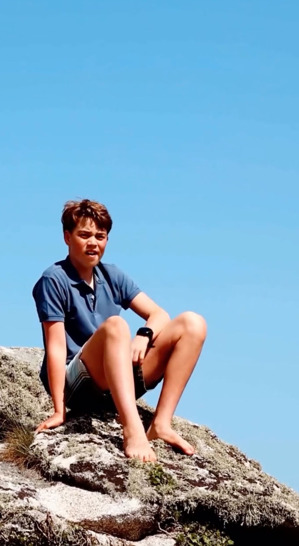 Prince George seated on a rock, wearing a blue polo shirt and shorts, looking at the camera.