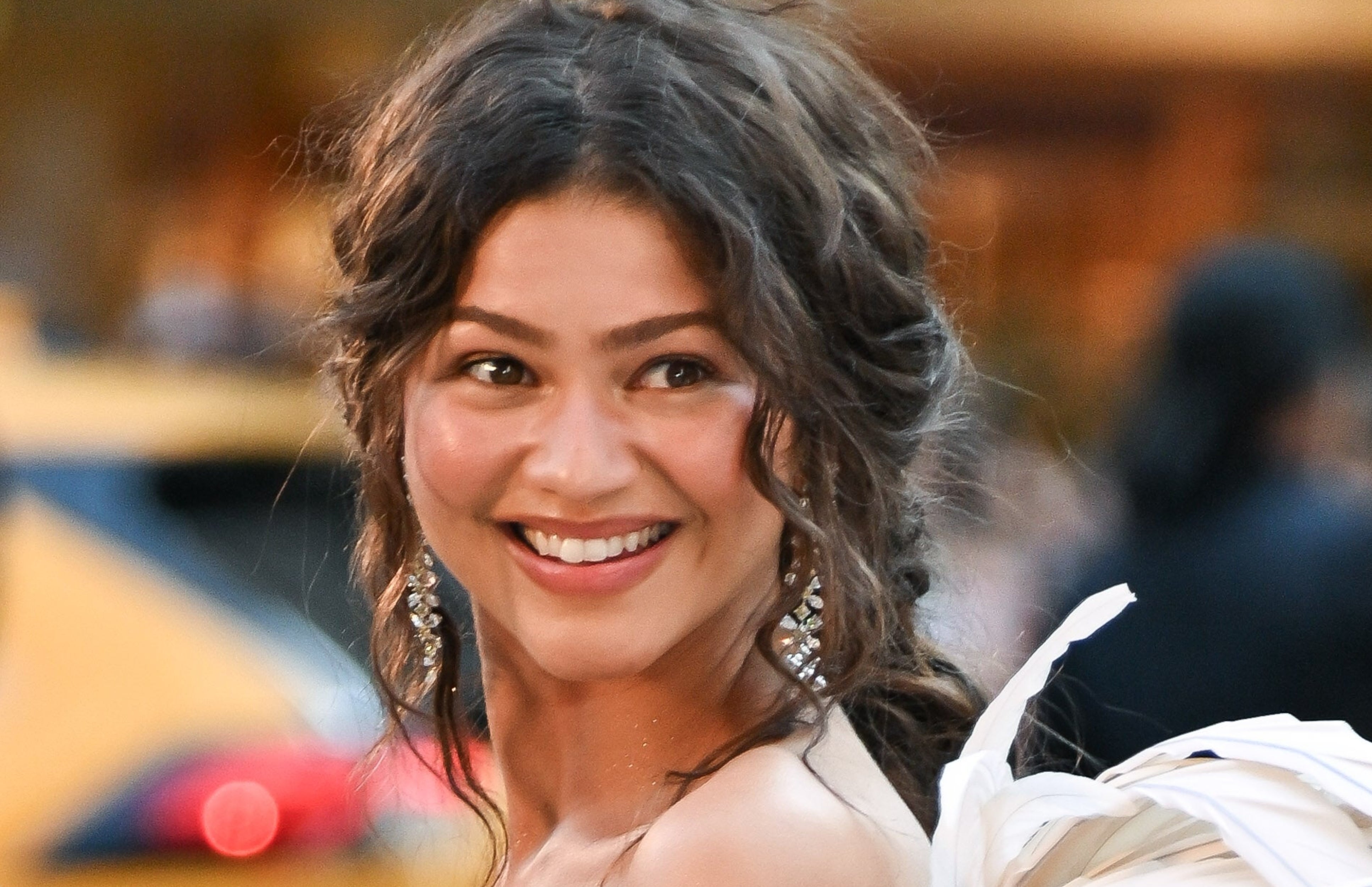 A person on the red carpet wears a strapless gown with feathered details. They're smiling as someone holds a can, possibly for a touch-up