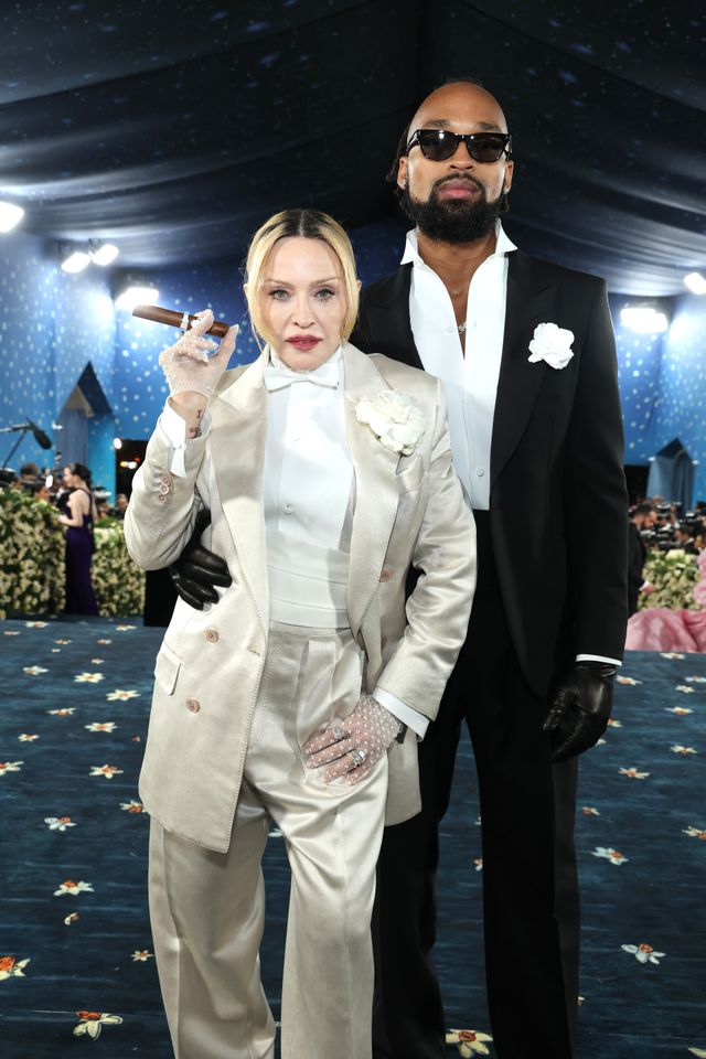 Two individuals in formal attire posing at an event one holding a cigar with decorative lights and floral arrangements in the background