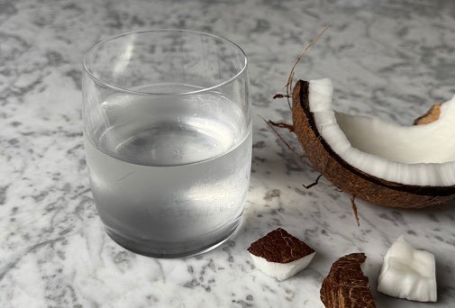 A glass of clear coconut water next to a cracked open coconut with pieces on a marble surface