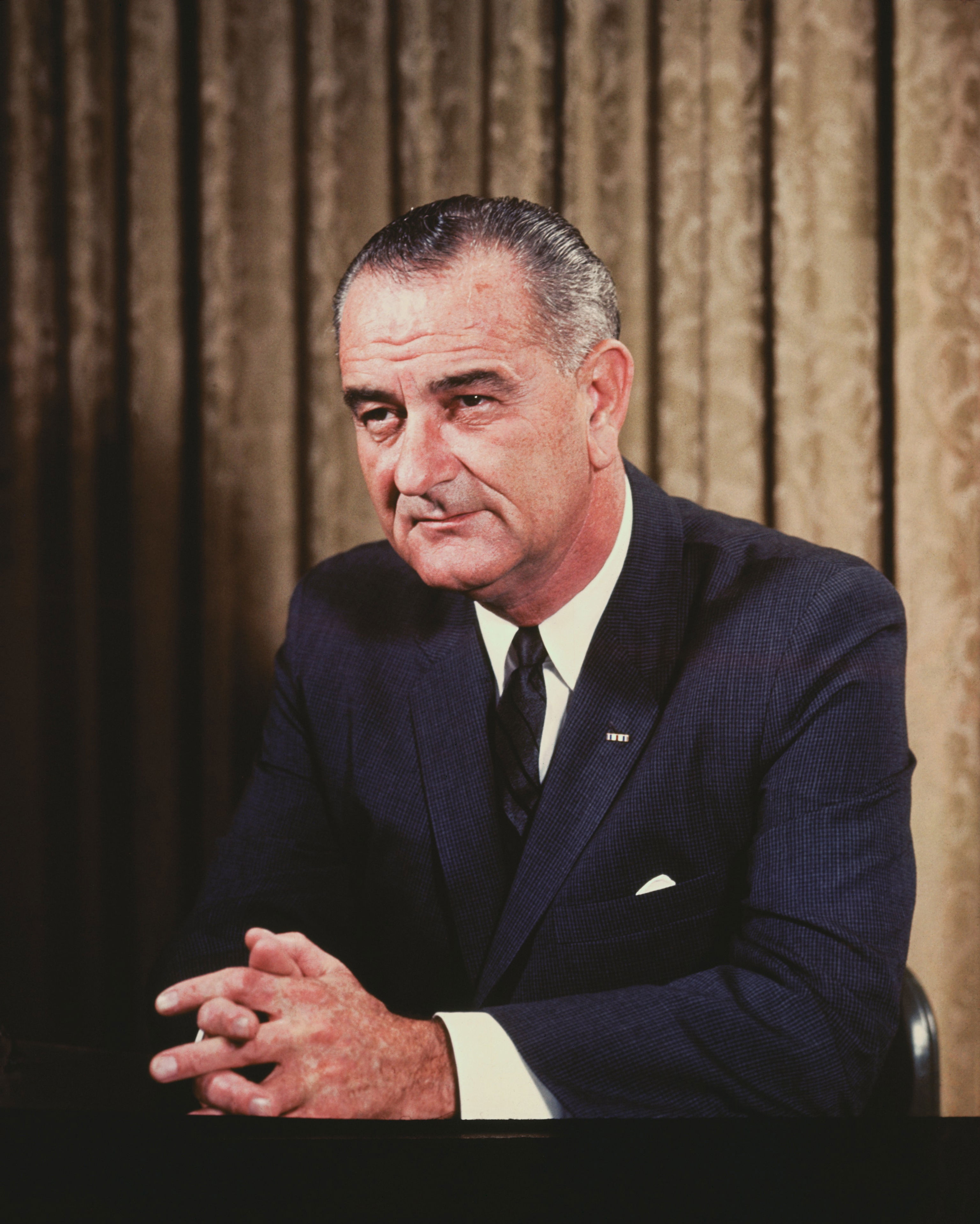 A distinguished man in a suit sits thoughtfully at a desk, hands clasped