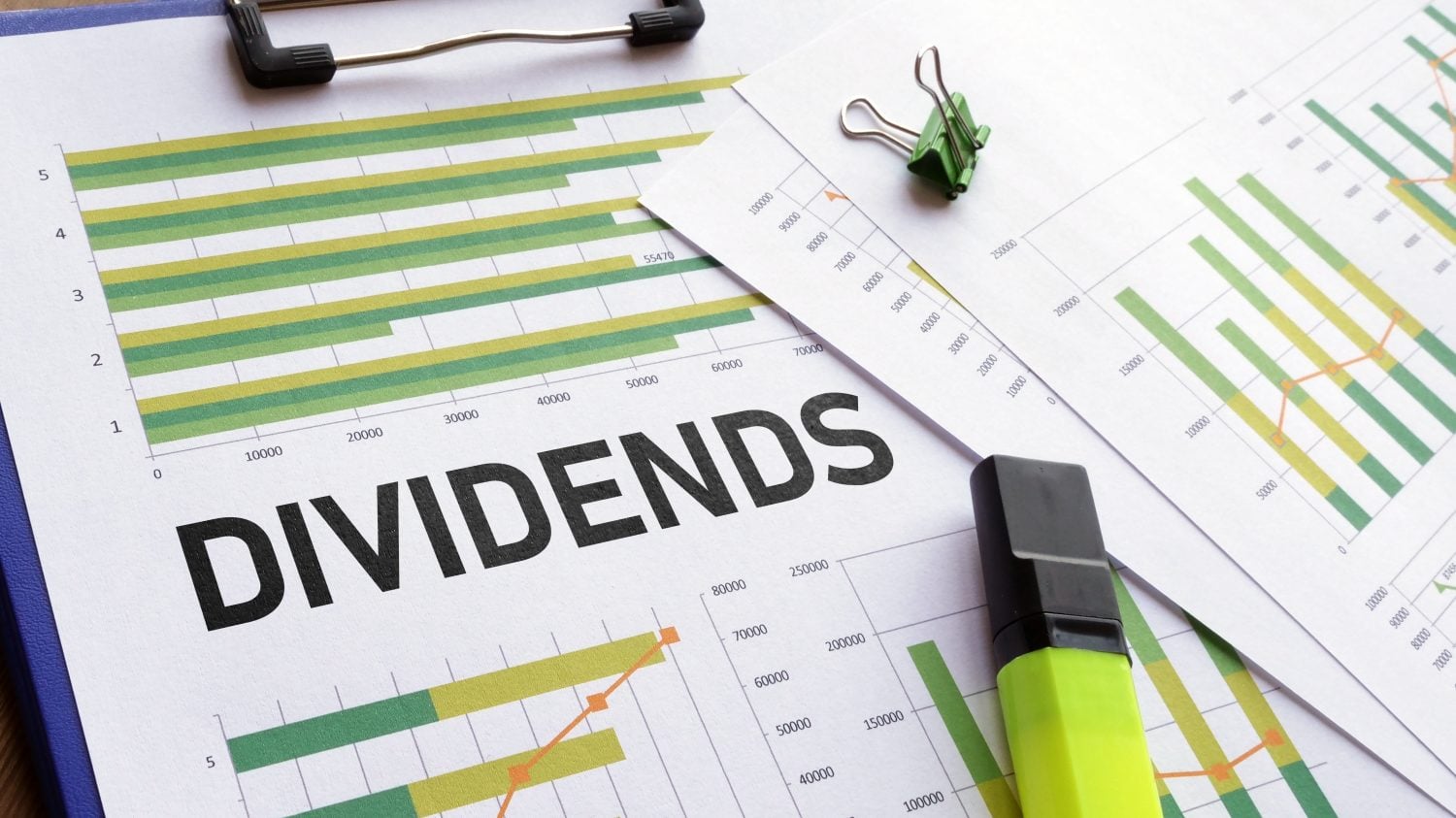 A flat lay photo shows multiple financial charts and graphs on white paper, held by a blue clipboard. The word 'DIVIDENDS' is printed in large black letters across the center of one sheet. A green binder clip secures papers at the top right, and a bright green highlighter rests in the lower right foreground. The charts feature horizontal and vertical bars in shades of green and yellow, with orange line graphs indicating trends and numerical scales along the axes.