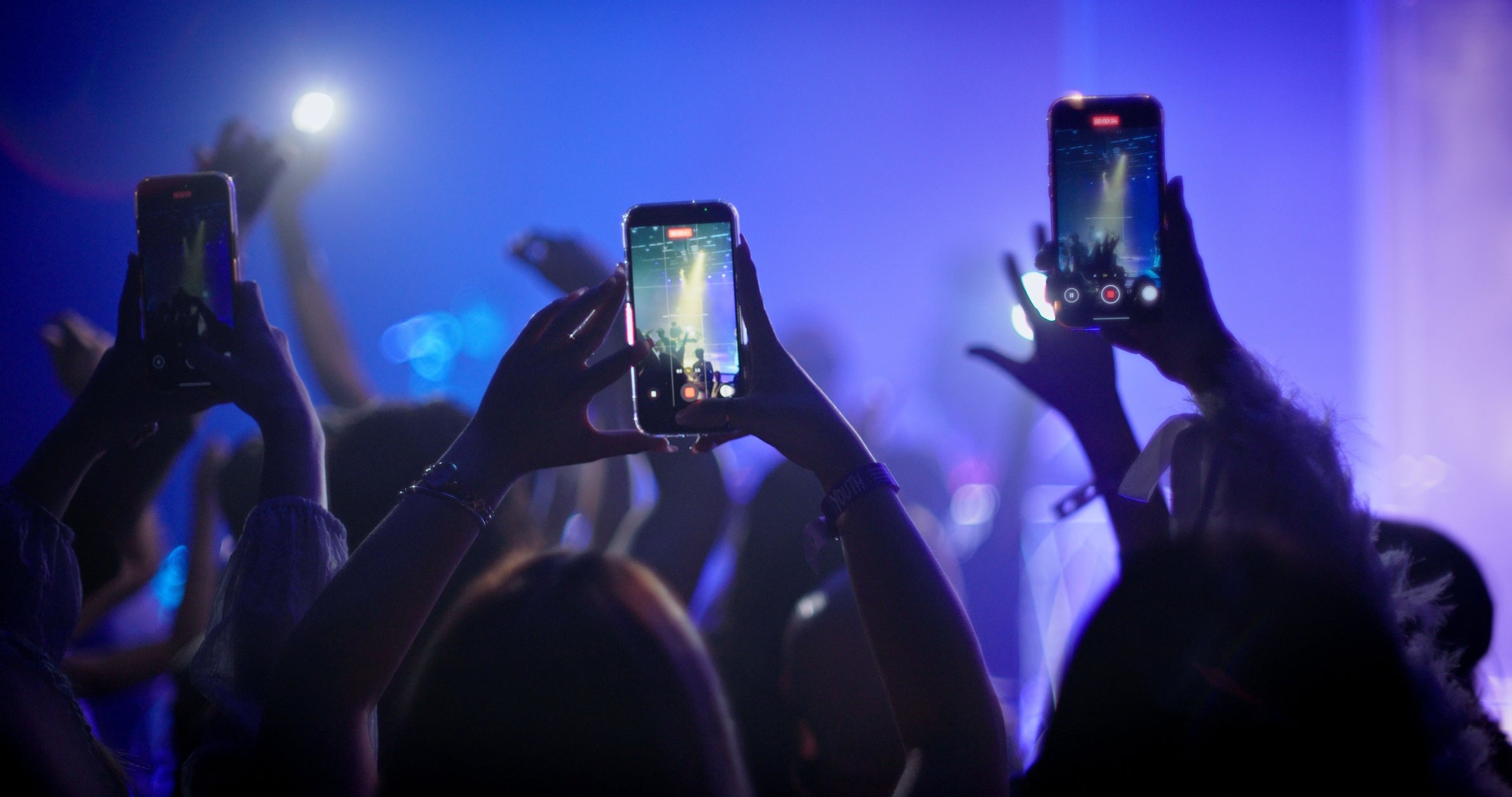 People at a concert holding up smartphones to record the performance, with a vibrant stage in the background