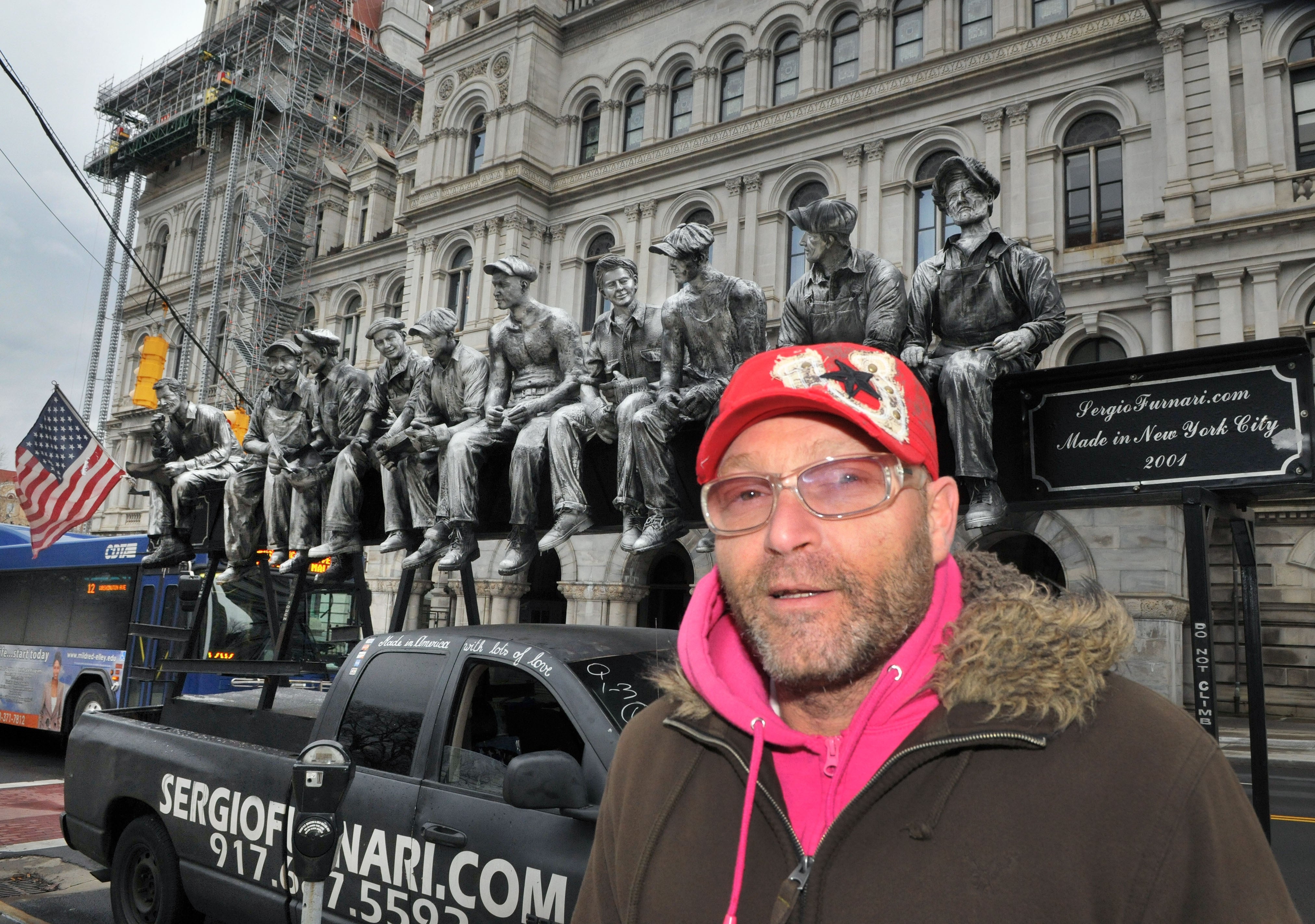Man in front of a truck with a sculpture of workers seated on a beam, similar to the famous "Lunch atop a Skyscraper" photo. Building in background