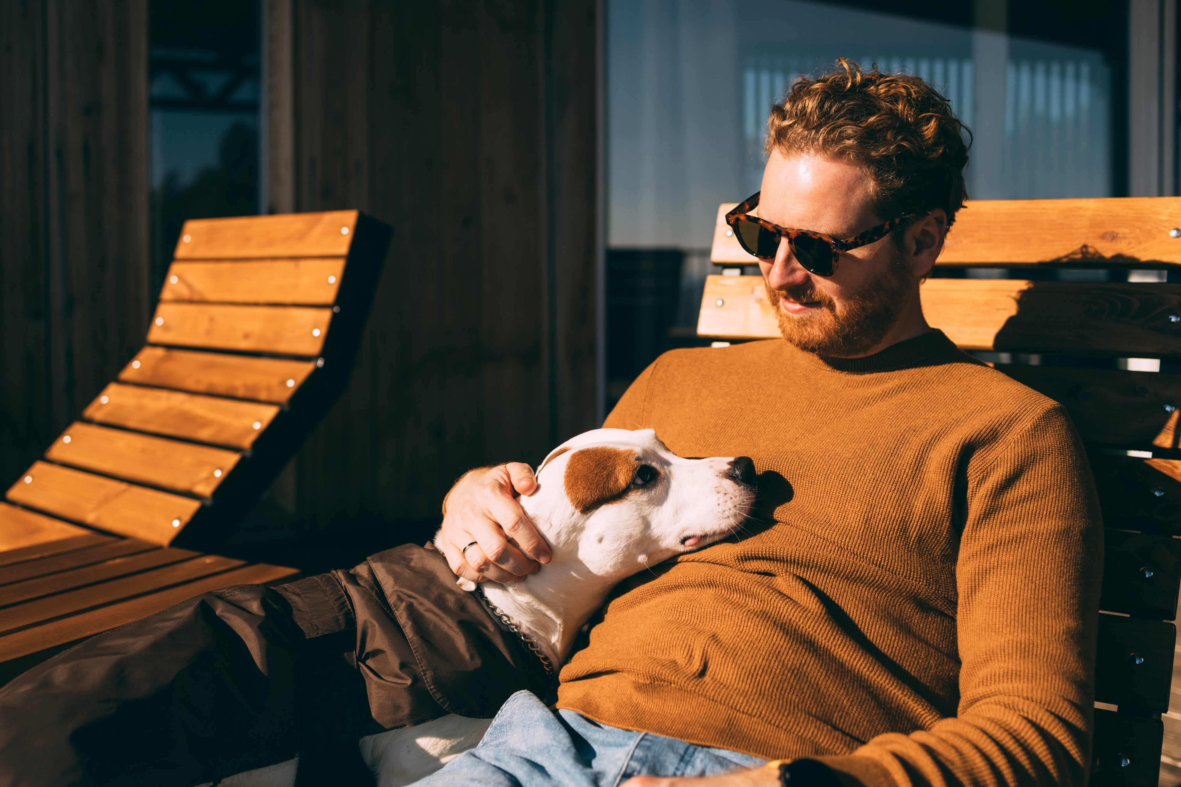 A man in casual sweater and sunglasses sits on a wooden lounge chair, affectionately petting a white dog resting on his lap