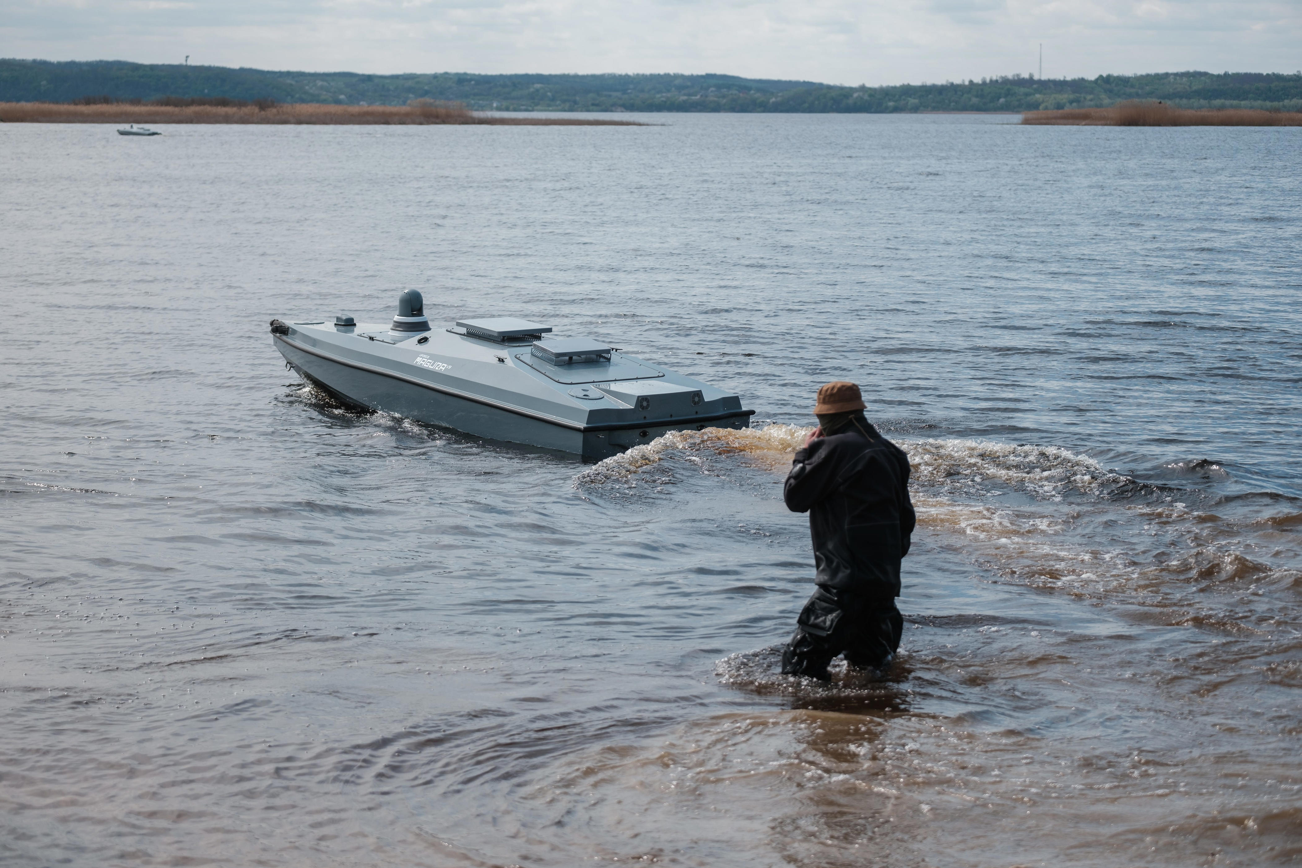 A person stands in shallow water near a small unmanned-looking boat moving across a wide river or lake.
